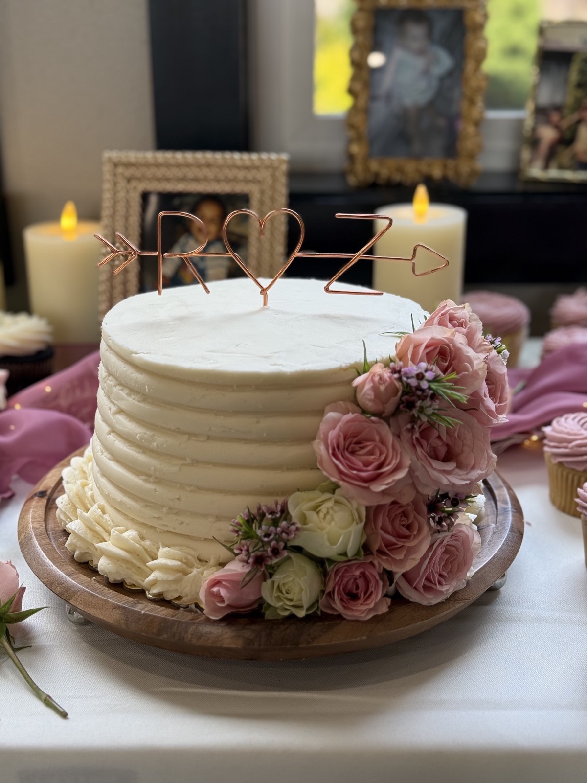 White buttercream celebration cake with pink roses and cupcakes nearby